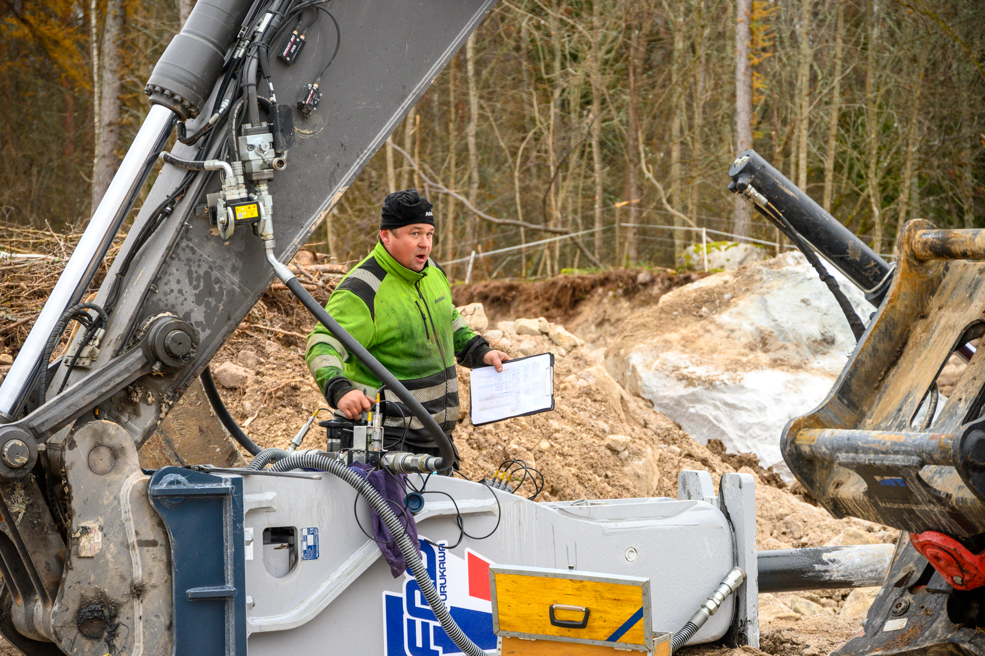 Arbetsledare med manual övervakar hydraulhammarfäste på grävmaskin vid anläggningsarbete utomhus