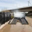 Mobil hjultvättstation i drift under öppen himmel med högtrycks vattendimma som rengör däcken på ett tungt fordon, placerad vid en byggarbetsplats med en bro och vindkraftverk i bakgrunden, vilket framhäver stationens miljövänlighet och effektivitet."