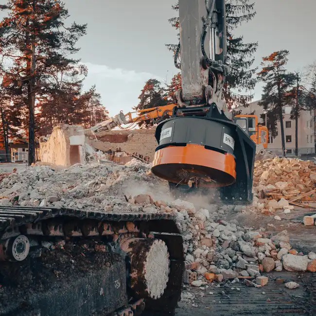 Sorteringsverktyg på grävmaskin hanterar återvinningsbart material vid en rivningsplats med betong och tegel i förgrunden och träd i bakgrunden