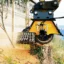 Närbild av en kraftfull stubbfräs i arbete, som skär genom en trädstubbe nära en träbyggnad. Maskinen producerar träflis och sågspån, omgiven av grönska och växtlighet.