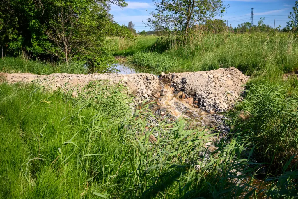 Vattenflöde vid Kölstaån under utbyggnaden av E18 omgiven av grön vegetation och grävningsarbeten