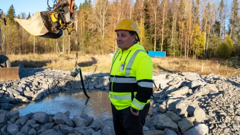 En byggarbetare i gul reflexjacka och hjälm står vid en vattenfylld arbetsplats medan en grävmaskin håller upp en dränkpump för att tömma vattnet. Skog i bakgrunden under en solig höstdag.