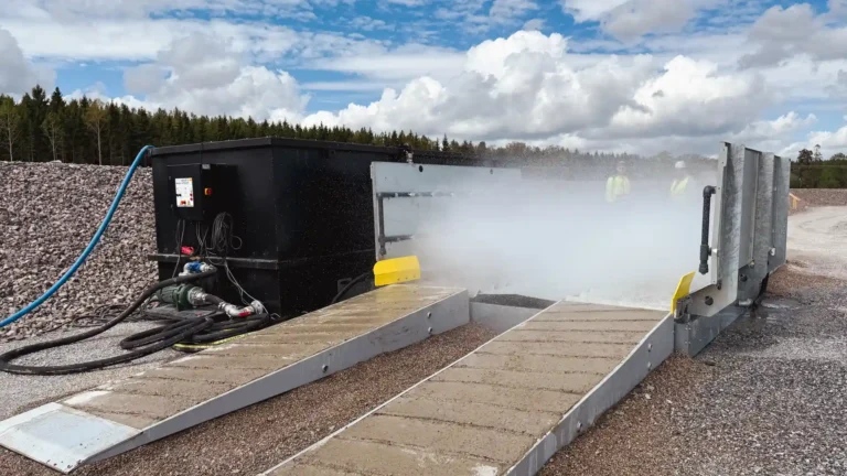 Mobil hjultvättstation i drift under öppen himmel med högtrycks vattendimma som rengör däcken på ett tungt fordon, placerad vid en byggarbetsplats med en bro och vindkraftverk i bakgrunden, vilket framhäver stationens miljövänlighet och effektivitet."