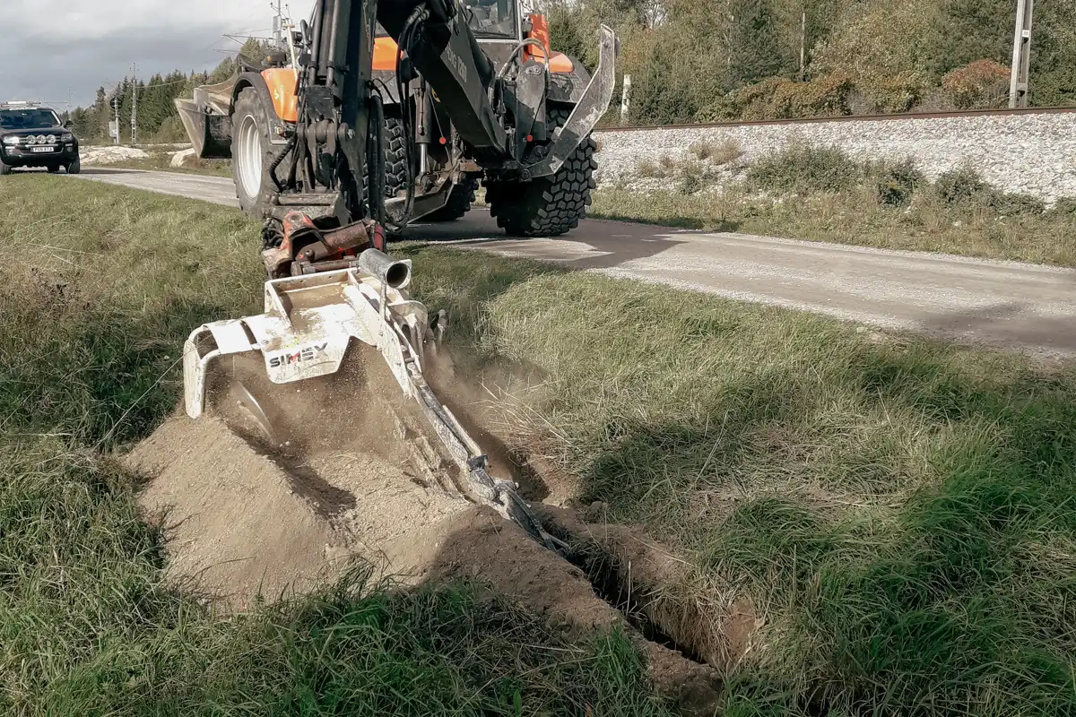 Simex kedjegrävare i arbete längs vägkant, där ett smalt och djupt dike grävs för installation av kablar eller dränering.