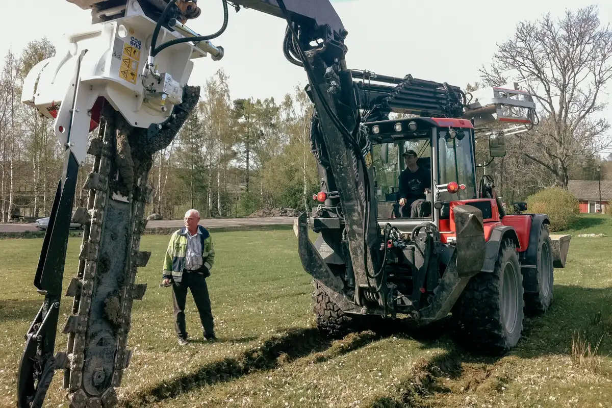 Simex kedjegrävare monterad på grävmaskin som gräver smalt dike i gräsyta för kabel- eller fiberdragning.
