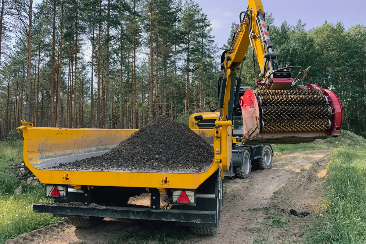 Grävmaskin med siktskopa lastar sorterad jord och massor på lastbil vid markarbete i skogsmiljö.