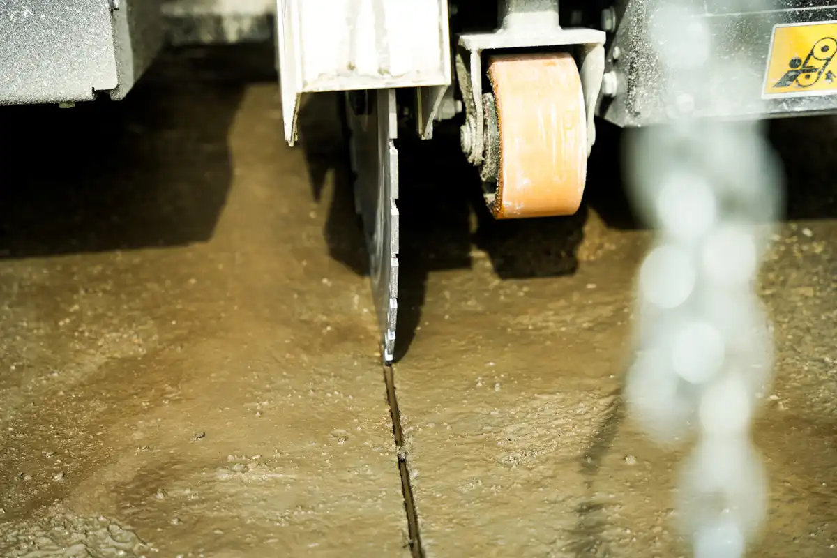 Concrete floor being cut by a floor saw; orange abrasive wheel and metal blade guard visible as the blade tracks a crack in the surface.